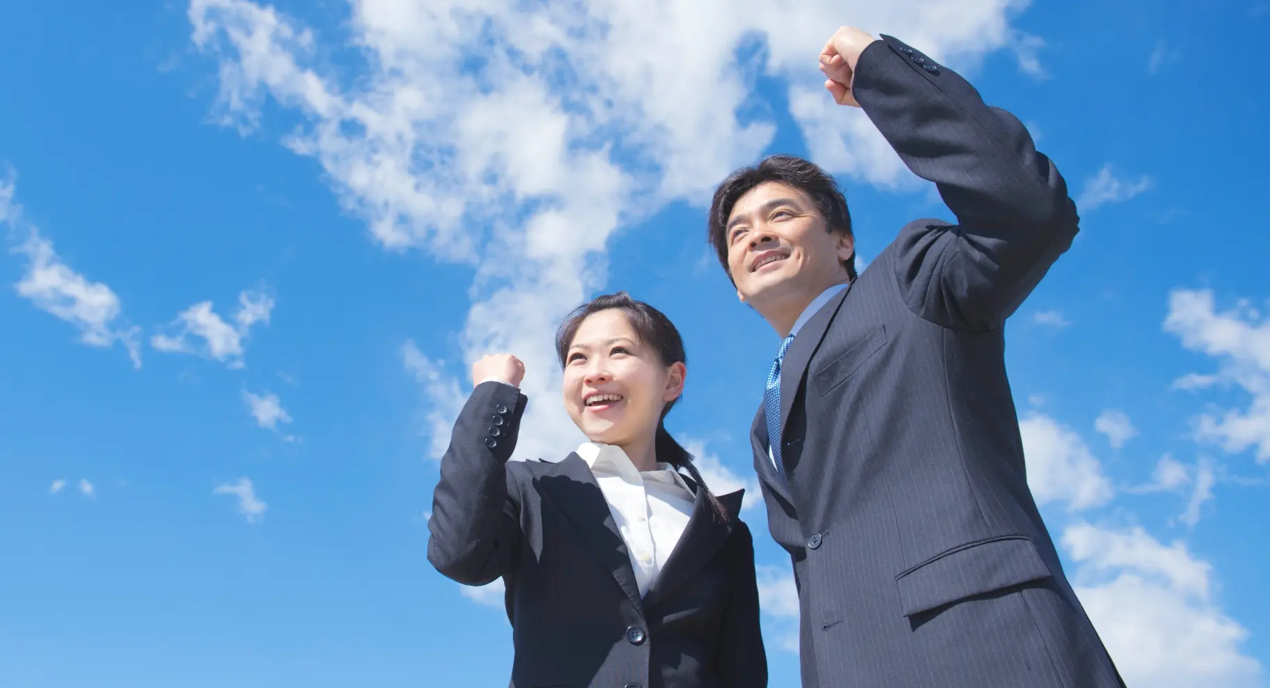 青空の下、スーツを着た男女が笑顔で拳を上げ、前向きな表情をしている