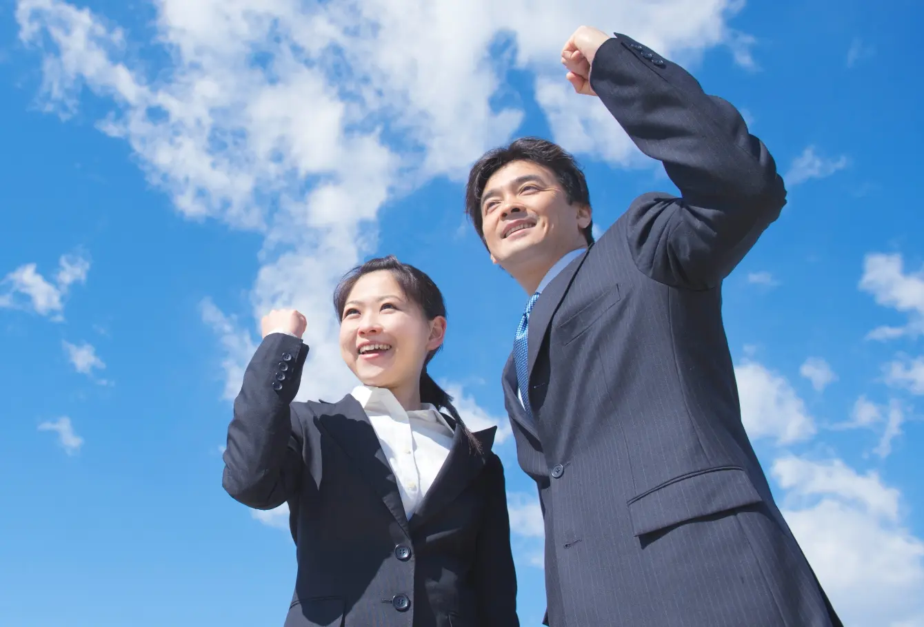 青空の下、スーツを着た男女が笑顔で拳を上げ、前向きな表情をしている｜採用情報イメージ